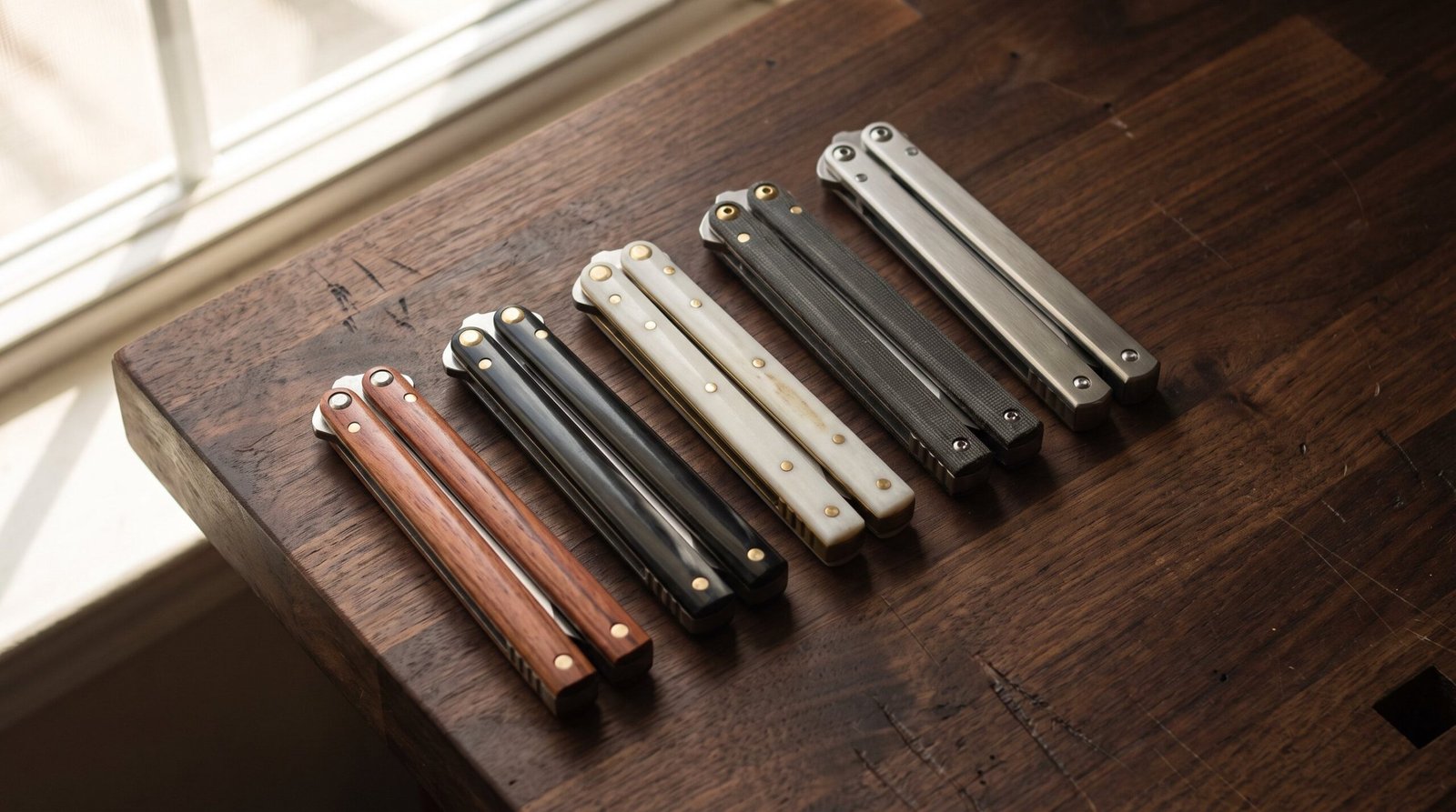 Handmade Filipino balisong butterfly knives lying closed in a row on a workbench, showing rosewood, buffalo horn, bone, micarta and stainless steel handles
