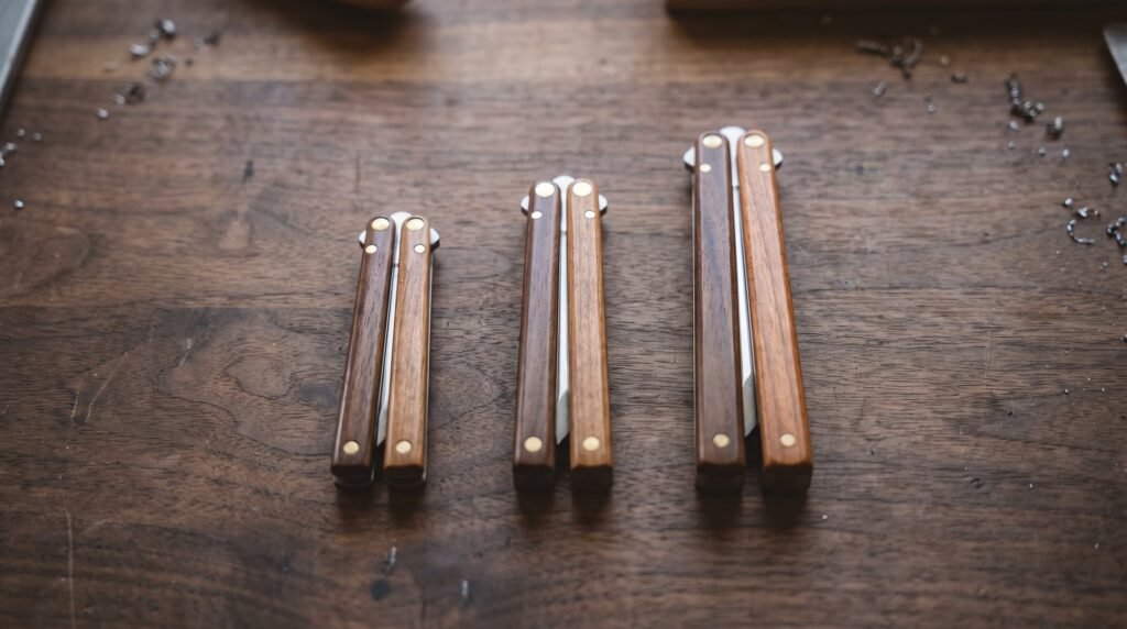 Three handmade balisong butterfly knives of different lengths lying closed side by side on a workbench