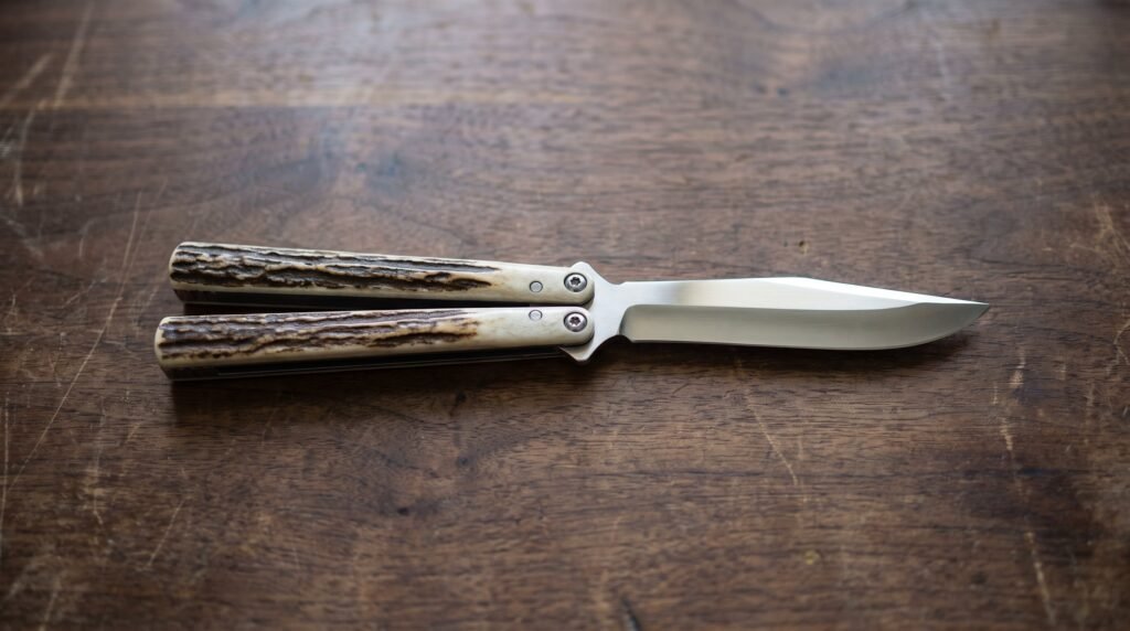 Single balisong butterfly knife lying fully open and flat on a dark walnut bench with stag handles and a polished satin blade