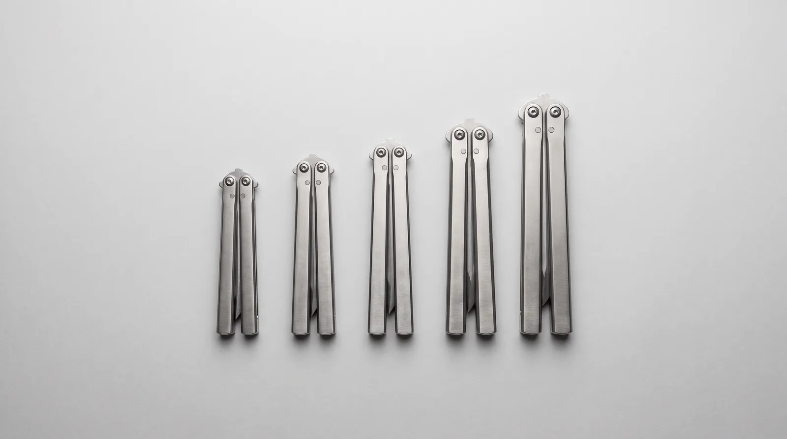 A row of five closed balisongs ascending in size on a light grey background