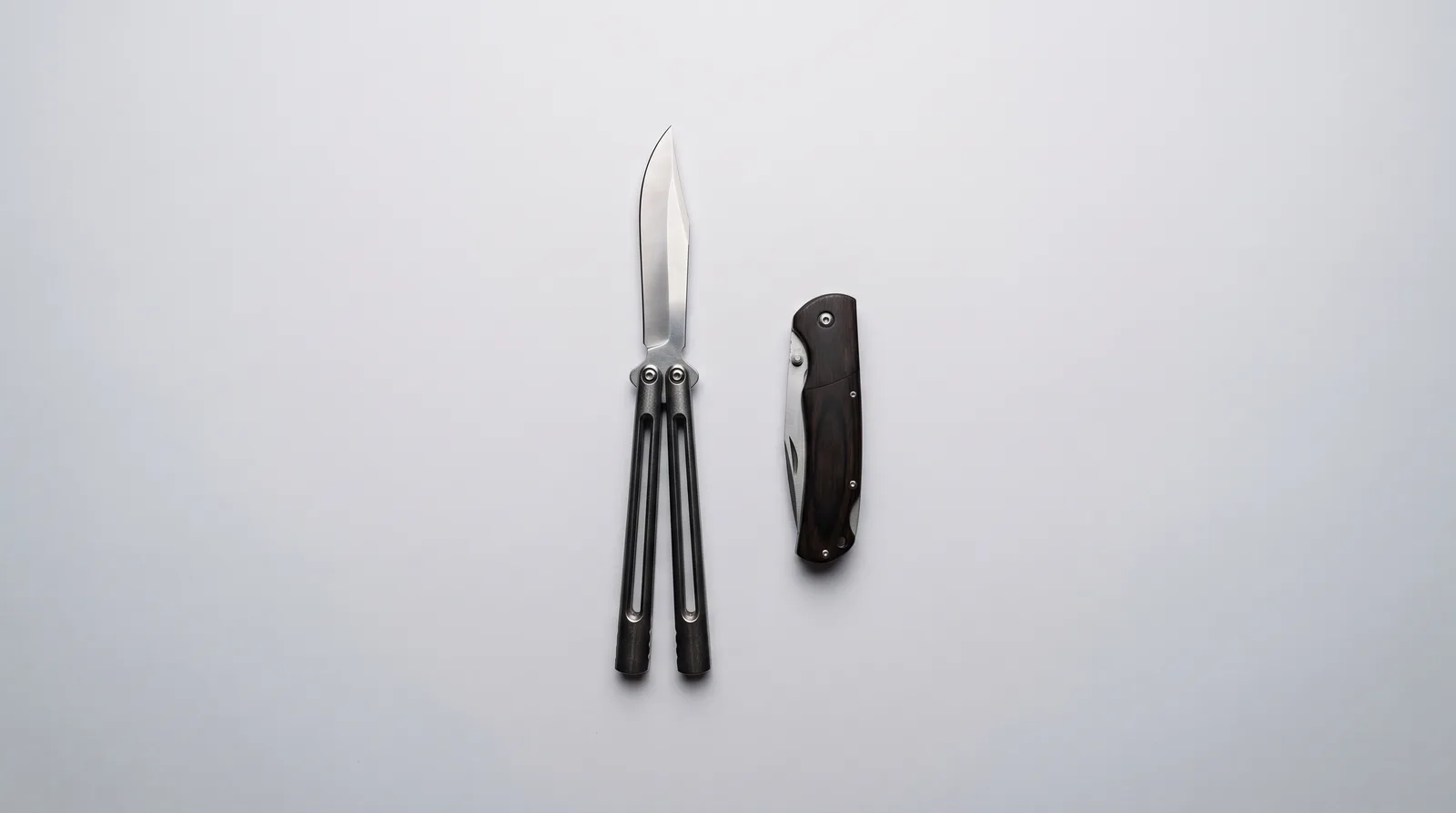 An open balisong and a closed folding pocket knife laid side by side on a grey background
