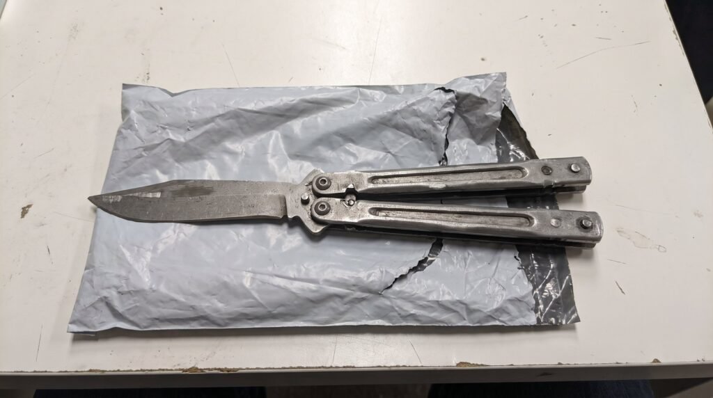 Cheap mass produced butterfly knife with a crude finish and uneven handle gaps lying on a plain shipping mailer