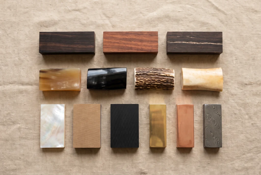 Balisong handle materials laid out on linen: kamagong, rosewood, wenge, carabao horn, black horn, stag antler, camel bone, pearl, micarta, G10, brass, copper and Damascus