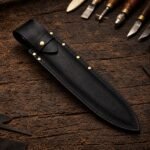 Handmade black full grain leather chef knife sheath with brass rivets and snap strap, lying on a wooden workbench