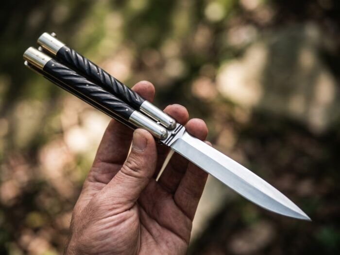 Mouse Filipino balisong butterfly knife held closed in hand outdoors with a blurred woodland background